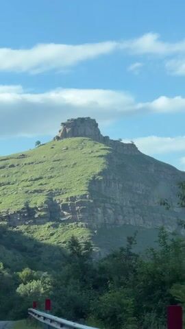 A video shows a rocky mountain peak covered in green grass under a blue sky with scattered clouds. The peak has a flat, layered top with some vegetation and what appears to be a small structure or flag on its summit. The slopes of the mountain are steep and show visible rock strata. In the foreground, the tops of trees are visible. The video appears to be shot from a moving vehicle, indicated by the slight blur and the presence of what looks like a red and white barrier at the very bottom of the frame.