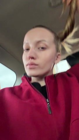 A woman with blonde hair pulled into a ponytail is sitting in the back seat of a car. She is wearing a red zippered jacket over a black top. She looks at the camera and briefly smiles. The car appears to be moving, and a yellow road sign is visible through the rear window.