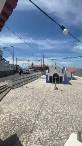 The video pans across a sunlit town square paved with cobblestones. In the center of the square stands a small, white chapel with blue trim and a red-tiled roof. A stone cross stands in front of the chapel. String lights with bare bulbs are strung across the top of the frame. To the left of the square, a street slopes downhill, with a few cars and people visible. Beyond the town, a distant view of the ocean and more buildings can be seen. The sky is blue with scattered white clouds.