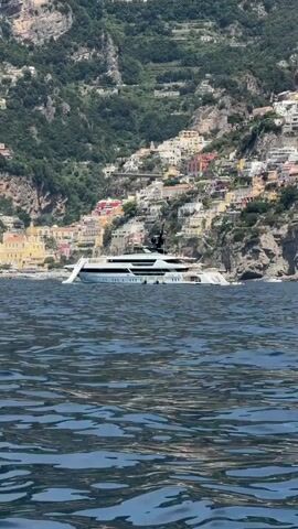 The video shows a large yacht in the ocean off the coast of a colorful town built into a mountain side. The water is a dark blue, with small waves reflecting the sunlight. A smaller white boat passes to the left of the yacht, obscuring it for a moment. The town is a mix of pastel colored buildings stacked on top of each other up the mountain side, and a light yellow church sits on the coast in front of the town. The mountain is covered in green trees.
