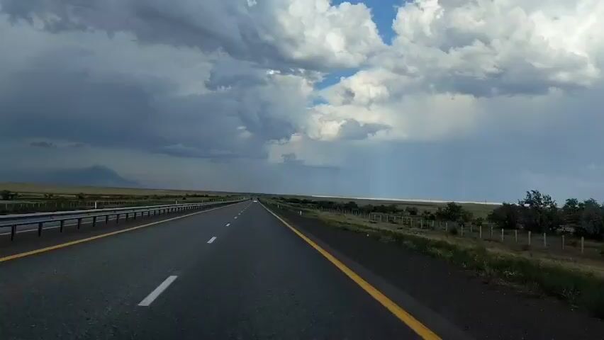 The video shows a first-person perspective of driving on a highway. The road is asphalt with a yellow line on the right and white dashed lines on the left. The sky is filled with dramatic, dark clouds, with patches of blue sky visible. Rain appears to be falling in the distance on the right side of the frame. There is a guardrail on the left side of the road and a fence with scattered trees on the right. In the far distance, mountains or hills are visible on the left horizon.