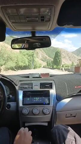 The video begins with a shot of the interior of a car, focusing on the dashboard and a person's hands on the steering wheel and gear shifter. The driver's side mirror shows a person in the distance, waving. The car appears to be moving along a road in a mountainous, rural area with trees and vegetation. The dashboard has a large screen with a green interface. The camera pans slightly to the right, revealing more of the car's interior and the scenery outside. The video ends with a continued shot of the road ahead and the surrounding landscape.