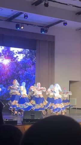 A group of women are performing a traditional dance on a stage. They are all wearing similar costumes: white blouses, blue skirts with colorful floral patterns, and headscarves. The women are arranged in a circle, moving rhythmically to the music. The stage has a wooden floor, and behind the dancers is a large screen displaying a video of a tree with purple flowers. There are stage lights above, and an audience can be seen in the foreground, watching the performance.