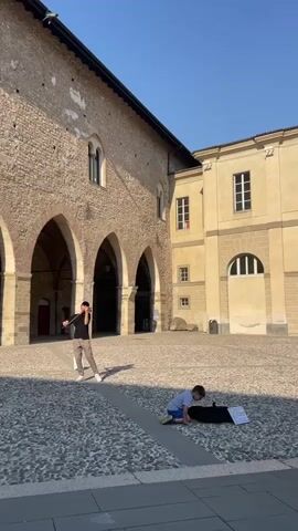 In an outdoor stone courtyard, a young adult stands playing the violin. He is wearing a black shirt, light brown pants, a black cap, and white shoes. A younger child sits on the ground nearby, possibly organizing something in a black case, with sheet music visible. The courtyard is surrounded by buildings with arched walkways and windows. The sky is clear and blue, and the scene is bathed in sunlight, creating shadows on the stone-paved ground. Another person approaches the violinist from the left side of the frame.