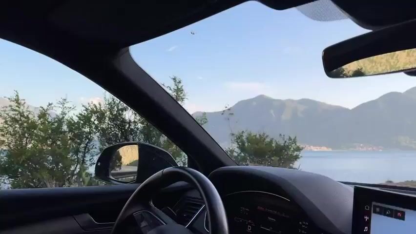 The video shows the view from inside a car as it drives along a road by a lake. We see the steering wheel, dashboard, and windshield of the car. The landscape visible through the windshield features a lake surrounded by mountains and trees under a clear sky. The car is moving, as evidenced by the changing scenery. Music plays in the background.