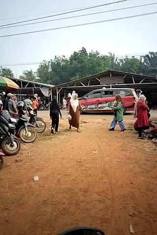 The video shows an outdoor market scene. In the foreground, the handlebars and part of the front of a motorcycle are visible, indicating the viewpoint is from a motorcycle. The ground is dirt. To the left, several motorcycles are parked. Behind the motorcycles, there are stalls or small buildings with roofs, and people are milling about. A woman wearing a brown dress and a white hijab is walking towards the camera, carrying a plastic bag in her right hand. She stops briefly and looks to her right. Further back, a red vehicle with graphics on its side is parked. More people are visible in the background, some walking and some standing near the stalls. The overall impression is of a busy market on a sunny day.