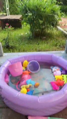 A low-angle, slightly shaky, vertical video opens on a purple inflatable kiddie pool filled with water and various colorful plastic toys, including a pink bucket, yellow duck, and several animal-shaped toys. The pool is situated on a wooden porch or deck, with a glimpse of a wooden bench and a lush green bush behind it. The background reveals a verdant garden setting with tall trees, suggesting a sunny day, possibly late afternoon due to the soft lighting and long shadows. The overall mood is peaceful and idyllic, evoking a sense of summer leisure and childhood fun. The video pans slightly upwards, showing more of the surrounding foliage and a white arched garden structure. There is no visible text in the media.