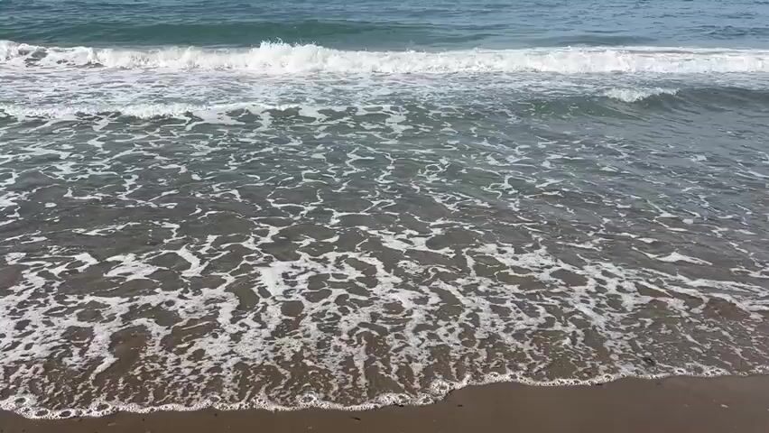 The video shows an ocean scene with waves gently rolling onto a sandy beach. The water is a deep blue, with white foam cresting the waves. The sand appears wet and dark where the waves have receded. There are no people or objects visible in the frame. The overall impression is one of a calm and natural beach environment.