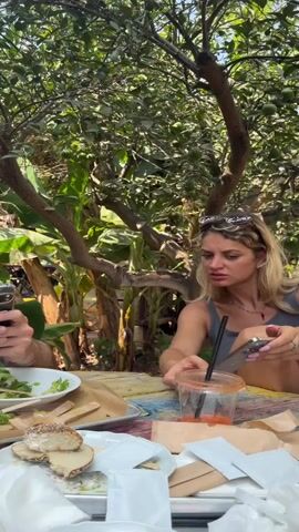 The video opens with a close-up of a woman with blonde hair, wearing a grey tank top and sunglasses pushed up on her head. She is looking down at her phone, which she holds in her right hand. In front of her is a table laden with food, including what appears to be a sandwich, a salad in a white bowl, and a clear plastic cup with a straw and orange liquid. To her left, part of another person's arm and hand is visible holding a fork, suggesting they are also eating. The background is filled with lush green foliage, with a tree bearing several small, unripe green fruits, possibly oranges or limes, prominently featured. Sunlight filters through the leaves, creating dappled light. The overall scene depicts an outdoor meal in a garden setting.