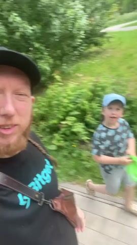 The video begins with a close-up selfie of a man wearing a black t-shirt with a logo, a black baseball cap, and a brown strap across his chest. He has a reddish-brown beard and a braid hanging from the back of his head. He is smiling and giving a peace sign with his left hand. The camera then pans slightly to the right, revealing a young child in the background walking on a paved path. The child is wearing a light blue baseball cap, a black t-shirt with a white pattern, and gray shorts. They are holding a bright green frisbee in their right hand. The environment appears to be a park or outdoor area with green grass and trees. The man continues to smile at the camera as he walks, with the child following behind him. The video then shifts to a slightly wider shot of the man, with the child still visible in the background, now further along the path and still holding the frisbee. The man then turns and walks away from the camera, and the video ends with a close-up of his face again, as he smiles and appears to be speaking. The child is no longer visible in the frame.
