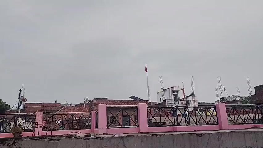 The video shows a rooftop view of buildings and the sky. A pink railing with black metal fencing runs across the foreground. Beyond the railing, there are several brick buildings, some with antennas and satellite dishes. A red flag is visible on a flagpole atop one of the buildings. The sky is overcast and gray. The overall scene suggests an urban or semi-urban environment under cloudy weather conditions. No people are clearly visible, and no specific actions or interactions are taking place.