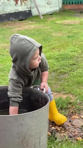 A young child wearing a green hooded sweatshirt and yellow rubber boots is playing with a large metal container filled with water. The child is holding a red elephant-shaped watering can and appears to be scooping water into it. The scene is outdoors, with green grass and a rustic building in the background. The child's face is partially obscured by the hood of the sweatshirt. The child's left hand is reaching into the water, and their right hand is holding the watering can. The watering can is tilted, and water is starting to spill out. The ground around the container is wet and appears to be muddy in some areas, with small stones and pebbles scattered about. The overall lighting suggests an overcast day.