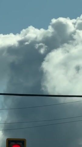 A low-angle shot captures a large, dark gray cloud with white edges against a bright blue sky. Below the cloud, dark power lines are stretched horizontally across the frame, with two thinner lines underneath. In the lower left corner, a traffic light shows a red signal within a yellow housing. The overall lighting suggests it is daytime.