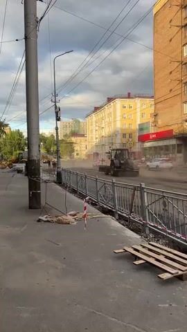 The video shows a construction site on a city street. A bulldozer is in the background, and workers are visible near some equipment. A fence is set up along the construction area, and there is dust in the air, suggesting ongoing work. Several cars are driving past the site on the street. The buildings in the background appear to be residential apartments.