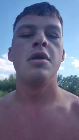 The video shows a shirtless young man walking along a paved road with trees and bushes along the side. He is holding a black piece of fabric, possibly a shirt or hat, in his left hand. He appears to be breathing heavily, and his face is somewhat flushed. The sky is blue with scattered clouds. The road curves slightly in the distance.