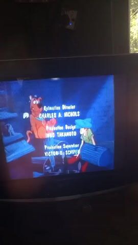 The camera shows a small television screen playing the end credits for a Scooby-Doo cartoon. The credits list "layout" personnel in blue text, with animated character portraits of Scooby Doo, Velma, Fred, and a skull, cycling through in sync with the credits. The room is dark, and the TV screen is the main source of light. A partially obscured hand is visible at the bottom left of the frame.