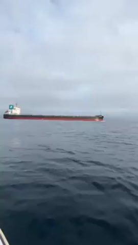 Here's a description of the video:

The video shows a large cargo ship moving across calm, dark blue water on a slightly overcast day. The ship is red and black, with a blue and white insignia on its front superstructure. The camera appears to be situated at sea level, possibly from a small boat, and the angle suggests the ship is moving from left to right in the frame. The sky is filled with grayish-white clouds. The focus is on the ship and its movement across the water.