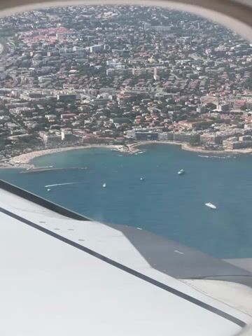 An aerial view from an airplane window shows a coastal city with a bay and a dense urban landscape extending inland. The clear blue water of the bay is dotted with several boats. The coastline features a beach area and buildings lining the shore. The city is densely packed with buildings of various sizes and colors, interspersed with trees. Hills and mountains form the background, disappearing into a hazy horizon. The edge of the airplane's wing is visible in the foreground, partially obscuring the view. The lighting suggests it is daytime, likely sunny.