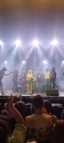 The video shows a musical performance on a stage. A female singer with blonde hair, wearing a colorful patterned dress, stands center stage holding a bouquet of pink flowers. She is surrounded by several male musicians, some holding instruments like guitars. Bright stage lights shine down from above, creating a hazy atmosphere. In the foreground, an audience watches, with some members raising their hands. The overall scene appears to be a concert or live event.