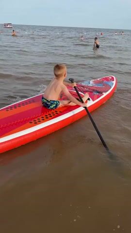 A young boy is paddleboarding on a body of water. He is sitting on a bright red paddleboard with orange and white accents. The paddleboard has a design of red and white abstract shapes. The boy is wearing colorful swim trunks and has blonde hair. He is holding a black paddle and is in the process of paddling, with water splashing from the paddle.

The scene is set on a body of water, likely a lake or the sea, given the horizon and other people visible in the distance. There are other individuals in the water, some of whom appear to be swimming or also paddleboarding. The sky is overcast, suggesting a cloudy day, possibly late afternoon or early evening. The water is a murky brown color.

The overall mood is one of outdoor recreation and enjoyment. The boy appears focused on his paddleboarding activity. The location appears to be a public beach or recreational area, with buoys marking a designated area in the water. The time of day is difficult to determine precisely due to the overcast conditions, but it seems to be daylight. There is no visible text in the media.