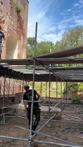 The video shows a construction worker in a green hard hat and black work clothes with a green logo on the back of his shirt working on a scaffolding structure. He is walking towards the left of the frame and appears to be adjusting or inspecting the scaffolding. The scaffolding is extensive, covering a large area with multiple levels of metal poles and planks. In the background, there is a partially visible brick building with an arched opening, suggesting it might be an old structure undergoing restoration. There are also trees and greenery visible behind the scaffolding. The ground is covered with debris and more scaffolding components, indicating an active construction site. The sky is visible with some clouds.