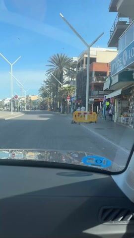 From the driver's seat, a vehicle moves along a street toward the ocean. The road is paved with gray blocks. Buildings line the street on the right. Palm trees and modern white light posts run along both sides of the street. Construction barriers stand near the right curb. The ocean and distant islands are visible in the background, with people walking and sitting along the water. The sky is blue and cloudless.