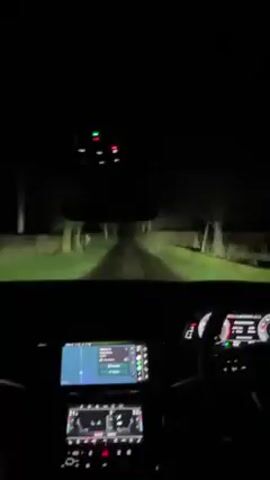 The video shows a car driving at night. The view is from the driver's seat, looking out the windshield. The road ahead is dark, but the car's headlights illuminate the road and the trees lining it. The dashboard is visible, with the navigation system showing a map. There are several buttons and dials on the center console. The car is moving forward on a dark, tree-lined road. The interior lights of the car are on. The car's headlights illuminate the path ahead. There is a navigation system on the dashboard. A hand is visible on the steering wheel.
