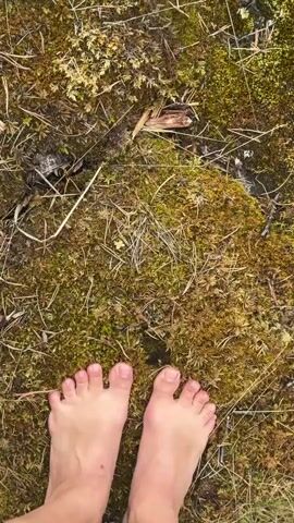 A close-up view of bare feet standing on mossy ground, then panning up to reveal a rocky cliff edge overlooking a body of water, with trees and a building visible in the distance under a cloudy sky.