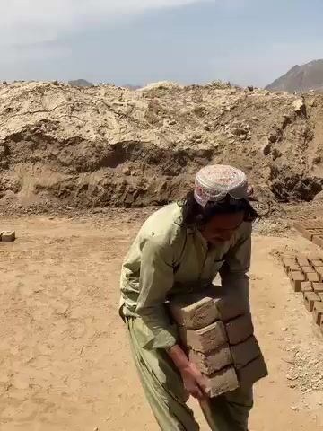 A man, dressed in a light green shalwar kameez and a knitted cap, is shown working barefoot in an arid, dusty outdoor environment. The setting features large, excavated dirt mounds in the background, with distant hills or mountains visible under a clear, bright sky. Rows of sun-dried mud bricks are laid out on the ground, stretching into the distance.

At the beginning of the video, the man is bent over, handling a stack of bricks near his feet, and picks one up. He then stands upright, makes a sweeping gesture with his hands, briefly looks at the camera with a smile, and then turns to walk away. Visible text on some of the bricks appears to read "BRICK".

The scene depicts a traditional brick-making site in Deh-E Bagh, Afghanistan, under a clear, sunny sky, likely during the daytime, given the strong shadows. The man's actions suggest a moment of demonstration, completion, or a brief interaction with the camera during his labor.