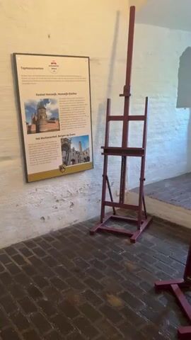 The camera pans across a small room, likely within a historical building or museum. The room features red brick flooring, white textured walls, and arches leading into adjacent spaces. There are two red easels positioned within the room. One is adjacent to an informational plaque, while the other is near the opening into the next space. A television monitor is mounted on one wall, and below it, there is an open wooden box with a screen inside. A decorative silver bench with a red cushion sits near the window. The room gives the impression of a historical setting with museum-like exhibits.