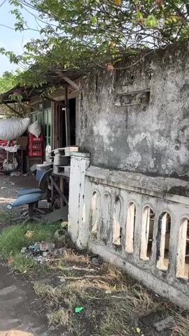 The video opens on a slightly elevated perspective looking at the exterior of a run-down building. The building's facade is a rough, weathered concrete with patches of moss and discoloration. To the right of the frame, a decorative concrete balustrade, also showing signs of age and wear, runs along the front of the structure. Overgrown vegetation, including vines and leaves, cascades from the roofline above.

The left side of the image reveals more of the building's exterior and its surroundings. A doorway, partially obscured, leads into what appears to be a small shop or dwelling. In front of the building, there are various objects scattered about, suggesting a state of disarray. These include a large white plastic bag, several red Coca-Cola crates, and what looks like a blue upholstered chair. There's also a wooden pallet stacked with items, and the ground is covered in debris, including plastic bags and scattered litter. The environment appears to be outdoors, with sunlight casting shadows and illuminating the scene, indicating it is daytime. The overall impression is one of a neglected, possibly impoverished, urban or semi-urban setting.
