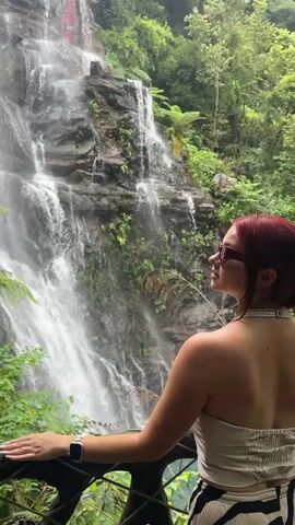 A woman with red hair is standing on a balcony, facing a waterfall. She is wearing a white strapless top and black and white striped pants. She is looking to her left, towards the waterfall. The waterfall is surrounded by lush green trees and foliage. The water is falling over a rocky cliff face. The balcony is made of wood and has a railing.