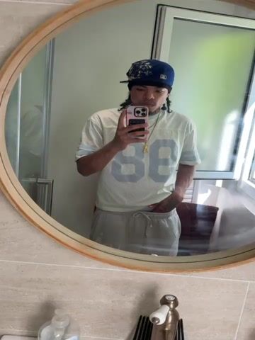 The media file features a young person taking a selfie in a bathroom mirror. The subject is wearing a light-colored jersey with the number "88" in a lighter shade, a blue baseball cap with a patterned bandana over the brim, and light grey sweatpants. A gold chain with a letter "A" pendant is visible around their neck. Their left hand is tucked into the waistband of their sweatpants, and their right hand holds a smartphone. In one frame, they are making a hand gesture with their right hand.

The setting appears to be a modern bathroom with light-colored tiled walls. A round wooden-framed mirror is the central object, reflecting the subject and the surrounding environment. In the reflection, a glass shower door, a white door, and a window with natural light streaming in are visible. The lighting suggests it is daytime. The overall mood is casual and relaxed, as indicated by the subject's attire and pose. There are no visible signs of specific weather conditions, but the natural light implies it is not dark outside.

The location context is Massy, France, although there are no explicit visual cues in the media file to confirm this specific location. The media file consists of a short video clip, presented as a series of still frames.
