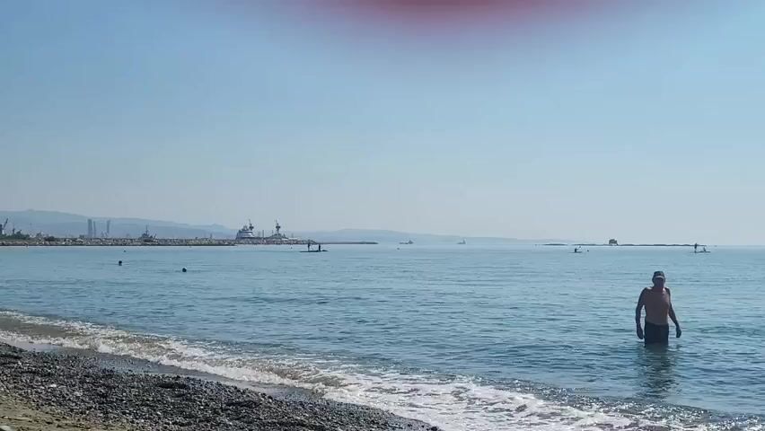 A video shows a beach with a person standing in the water. The person is wearing a hat and swimming trunks. The water is blue and calm, and there are waves lapping at the shore. The sky is clear and blue, and there are some clouds in the distance. There are also some boats in the distance, and a pier. The beach is made of pebbles. A red smudge is visible in the top center of the frame.
