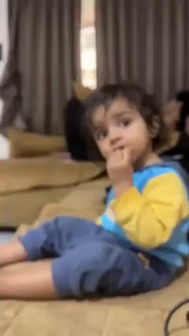 A young child is sitting on a beige couch, eating. The child is wearing a blue and yellow hooded sweatshirt and blue pants. Their hair is dark and curly. Behind them, another person is lying on the couch. The room has brown curtains and beige walls. A door is visible in the background. The child stops eating, looks to the side, and waves with their left hand.