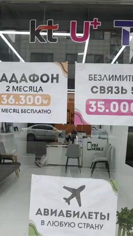 A view through a store window shows advertisements for mobile phone plans and plane tickets. The store name "KIM MOBILE" is visible above the window. Inside the store, a person is sitting at a counter. The advertisements are in Russian and promote unlimited 5G service for 35,000 won and plane tickets to any country. A security camera is mounted on the exterior of the building.