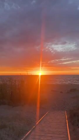 The video shows a sunset over a beach with a wooden boardwalk leading towards the water. The sun is a bright orange disk on the horizon, emitting a strong glare. The sky is colored with shades of orange, pink, and gray clouds. Beach grass grows on either side of the boardwalk on the sandy beach. The ocean appears calm.