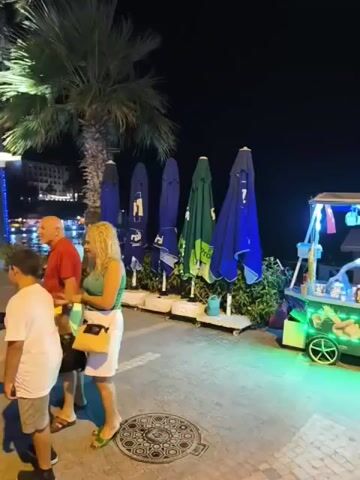 This video shows a nighttime scene in an outdoor area, likely a promenade or boardwalk. In the foreground, there are several people walking and standing. To the left, a couple stands near a row of closed umbrellas. A woman with blonde hair and a green shirt is facing away from the camera, while a man in a red shirt is partially visible. Further to the right, a food cart is illuminated with neon lights, displaying the word "MISIR". A person is operating the cart, and a woman in a black dress walks past it. Two men in casual attire walk by the food cart. In the background, palm trees are visible against the dark night sky. The ground is paved with cobblestones and has a patterned manhole cover. The overall ambiance suggests a relaxed evening setting, possibly a tourist area.