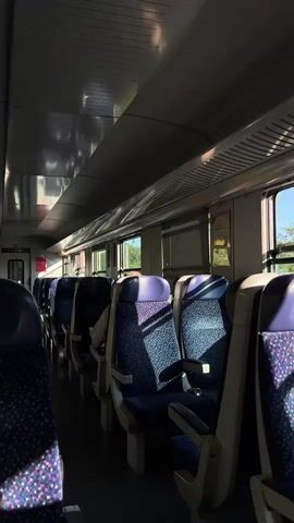 The video shows the interior of a train carriage with rows of empty seats. The seats are upholstered in a dark blue fabric with a speckled pattern of pink and white dots, and have purple headrests. Sunlight streams in through the windows on the right side of the carriage, casting shadows on the seats. The overall impression is one of stillness and calm, with a few passengers visible in the distance. The lighting suggests it is daytime.