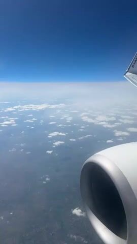 The video shows a view from an airplane window. The sky is a clear blue, transitioning to a hazy white horizon. Below the clouds are scattered across a visible landscape. In the foreground, a portion of the airplane's engine is visible. There are no people visible.