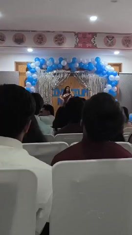A young woman stands on a stage, holding a microphone and singing. She is wearing a maroon dress and stands behind a podium with a banner that reads "CONVOCATION". The stage is decorated with blue and white balloons and silver streamers. An audience of people sits in chairs facing the stage. The room appears to be an event hall, with a white ceiling and decorated walls.