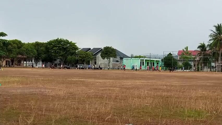 The video shows a dry, brown grassy field with people playing soccer in the distance. There are buildings and trees in the background, and the sky is overcast. Some people are running, kicking a ball, and generally engaging in activities related to playing soccer. The overall scene depicts an outdoor recreational activity.