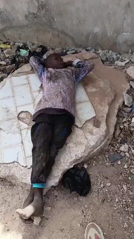A young boy lies on his back on a broken slab of tile and concrete rubble in what appears to be an alley or derelict lot. He is wearing a light-colored long-sleeved shirt with a zebra-like pattern, black pants that are dirty and possibly torn, and a blue band around one ankle. His hands are behind his head, and he seems to be asleep or resting. The area is littered with debris, including stones, plastic bags, and pieces of broken tiles. A black plastic bag sits near his legs. The environment suggests poverty or neglect.