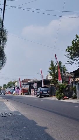 The video captures a roadside scene from a low-angle perspective. The ground is paved with asphalt, showing some wear and tear. To the left, a grassy verge is visible. Along the right side of the road, a row of small businesses or stalls are set up, some appearing to be shops or food vendors. Several Indonesian flags, red and white, are prominently displayed, attached to poles and fluttering in the breeze. A dark-colored car is parked on the side of the road near one of the stalls. Palm trees and other greenery are interspersed among the buildings. Overhead, power lines crisscross the bright, partly cloudy sky. The overall impression is that of a typical street scene in a tropical or subtropical location, possibly decorated for a national celebration.
