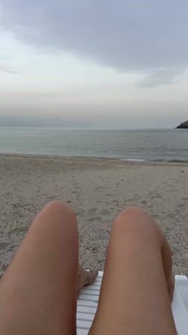 The video shows a person lying on a white plastic beach chair on a sandy beach, looking out at a body of water with a small rocky island visible in the distance. The camera is positioned from the perspective of the person on the chair, with their legs and feet visible in the foreground. Their feet are covered in sand. The sky is overcast, and the water appears calm. The person speaks and mentions, “There is a turtle beach in Fujairah. Oh, my dirty legs.”