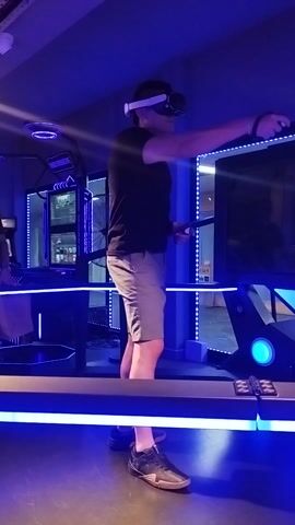 A man is playing a virtual reality game in an indoor, dimly lit setting, likely an entertainment center or arcade in Punta del Este, Uruguay. He wears a white VR headset and holds two controllers, actively moving his hands as he interacts with the virtual environment. He is dressed in a black t-shirt, khaki shorts, and dark sneakers.

The area is illuminated primarily by prominent blue LED strip lights that outline various structures and provide ambient light, contributing to a futuristic or immersive atmosphere. In the background, a large overhead screen reflects parts of the room and other digital displays, but no specific game content is clearly visible on it. The man appears focused and engaged in the game. No discernible text is visible in the scene.