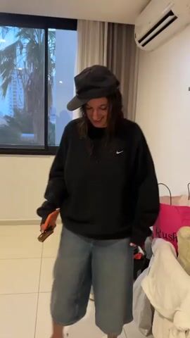 The video features a person, likely female, standing in an indoor setting, appearing cheerful and playful. She wears a black baseball cap (worn backwards), a black Nike sweatshirt with a white swoosh logo, and light blue, baggy denim shorts or capris. She holds an orange object, possibly a phone or remote, in her right hand and is seen smiling and moving rhythmically, suggesting she might be dancing or swaying to music.

The background includes a large window through which palm trees and distant buildings are visible, indicating a sunny, possibly warm climate. A white air conditioning unit is mounted on the wall above the window. To the right of the person, a pink shopping bag with the word "RUSH" printed on it and a pile of clothes are on the white tiled floor. The room has white walls and a white tiled floor. The scene depicts a relaxed and joyful moment, set in Dakar, Senegal.