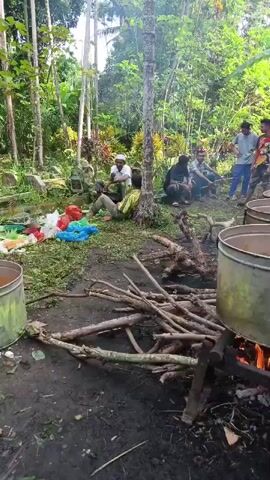 The video shows a group of people gathered outdoors in a wooded area. In the foreground, a fire is burning beneath two large metal pots, with a pile of firewood next to it. To the left, several colorful plastic bags are on the ground, along with some green foliage. In the middle ground, several men are sitting or crouching. One man in a white shirt and cap is talking to another man wearing a yellow shirt, who is leaning against a tree. Another man in a black shirt is seated behind them. In the background, more trees and foliage are visible, along with what appear to be gravestones or monuments. Several men stand in the far right of the frame. The overall scene suggests an outdoor gathering, possibly a communal cooking event or a funeral.