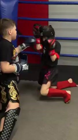 Two children are engaged in a sparring match inside a boxing ring. The ring has blue and red padded corners and white rope boundaries. One child, presumably a girl, is kneeling and wearing a black headgear, black and red boxing gloves, a black shirt with red accents, black shorts with pink designs, and red shin guards. She is actively throwing punches at the other child. The second child, presumably a boy, is also kneeling and is wearing black and white boxing gloves, a black t-shirt with a colorful graphic on it, and black and gold shorts. He also has on black and white shin guards. The boy is actively blocking the punches thrown by the girl. The floor of the ring is gray.