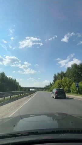 The video shows a view from inside a car driving down a two-lane road on a sunny day. The sky is blue with scattered white clouds. Trees and vegetation line the sides of the road, and a guardrail is visible on the left. In the distance, an overpass or bridge can be seen. A dark-colored SUV is driving in the same direction on the right lane, ahead of the car from which the video is being filmed. The dashboard of the filming car is visible in the foreground.